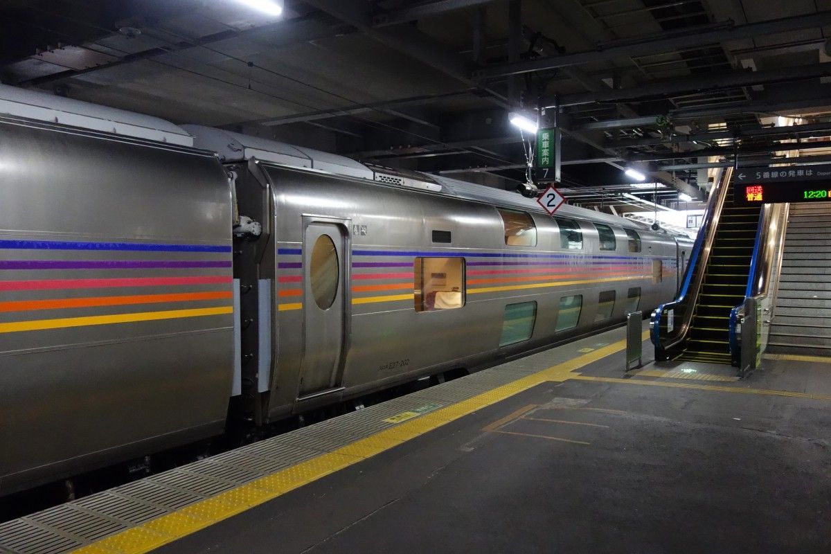 Bare brushed metal train cars with rounded edges, bright  blue, purple, pink, orange and yellow thin stripes painted all along, waiting at a platform with yellow safety paintings, with an escalator with blue handrails. Part of the red, green and orange LED direction sign is visible.