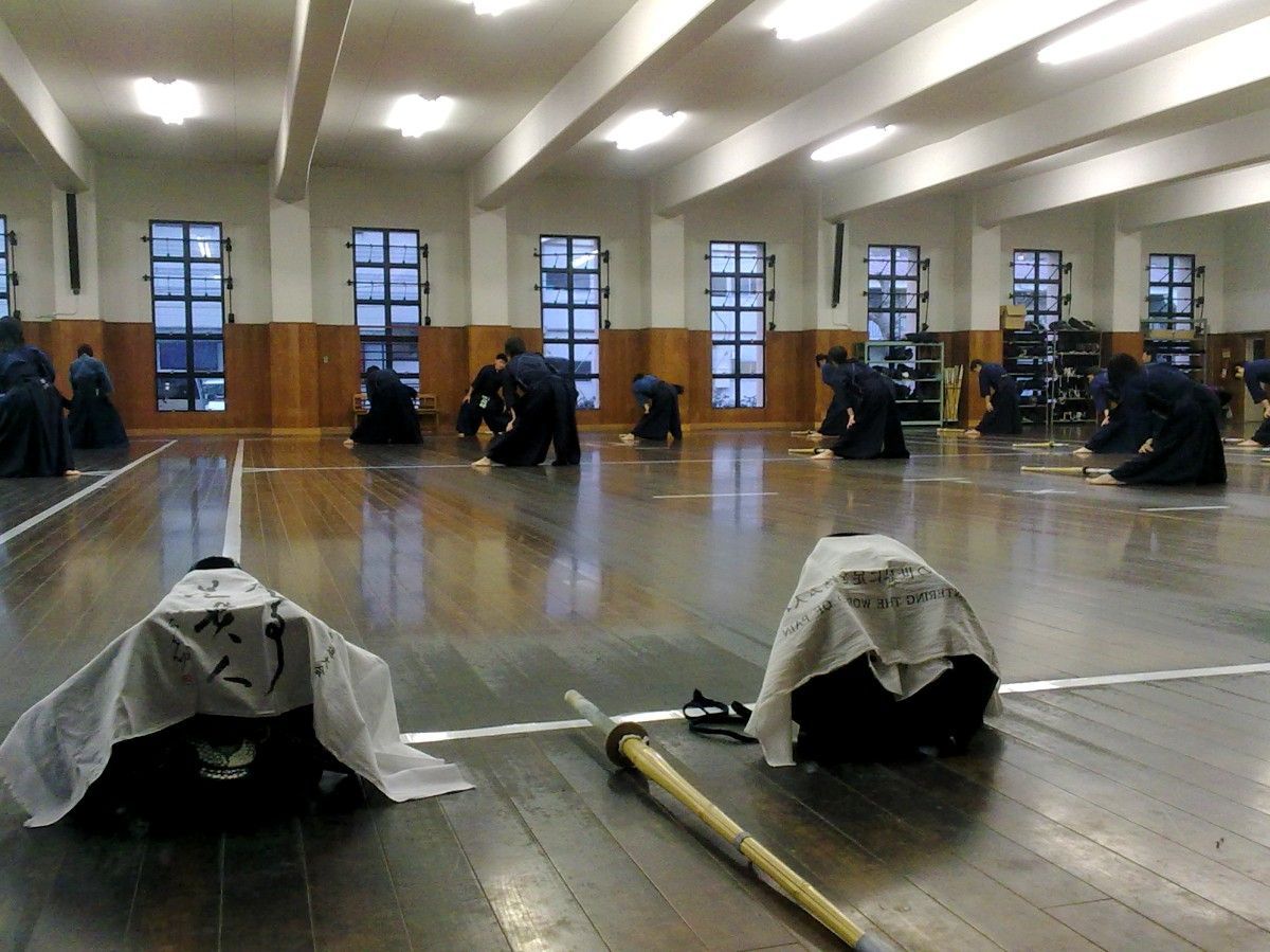 several students in dark blue kendo attire (hakama and vest, with pieces of armor) are lined up on the wooden flooring are stretching
2 helmets covered with towels are sitting on the foreground. One bamboo sword lays between them