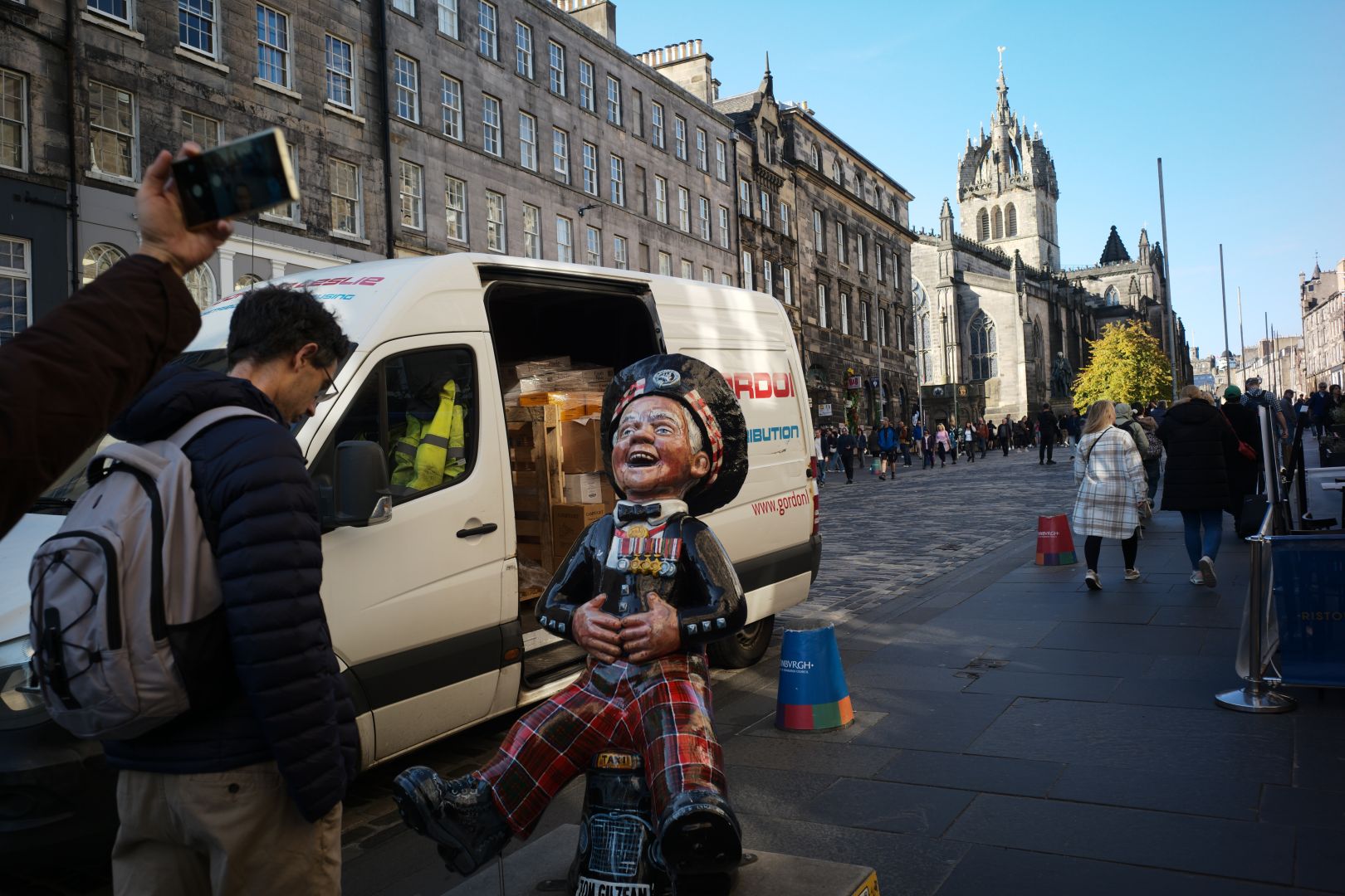 Street view with a guy effigy leaning backwards laughing. Behind it is a white van and an unsuspecting tourist and another disembodied hand taking a phone photo. I should google what that little status is about….