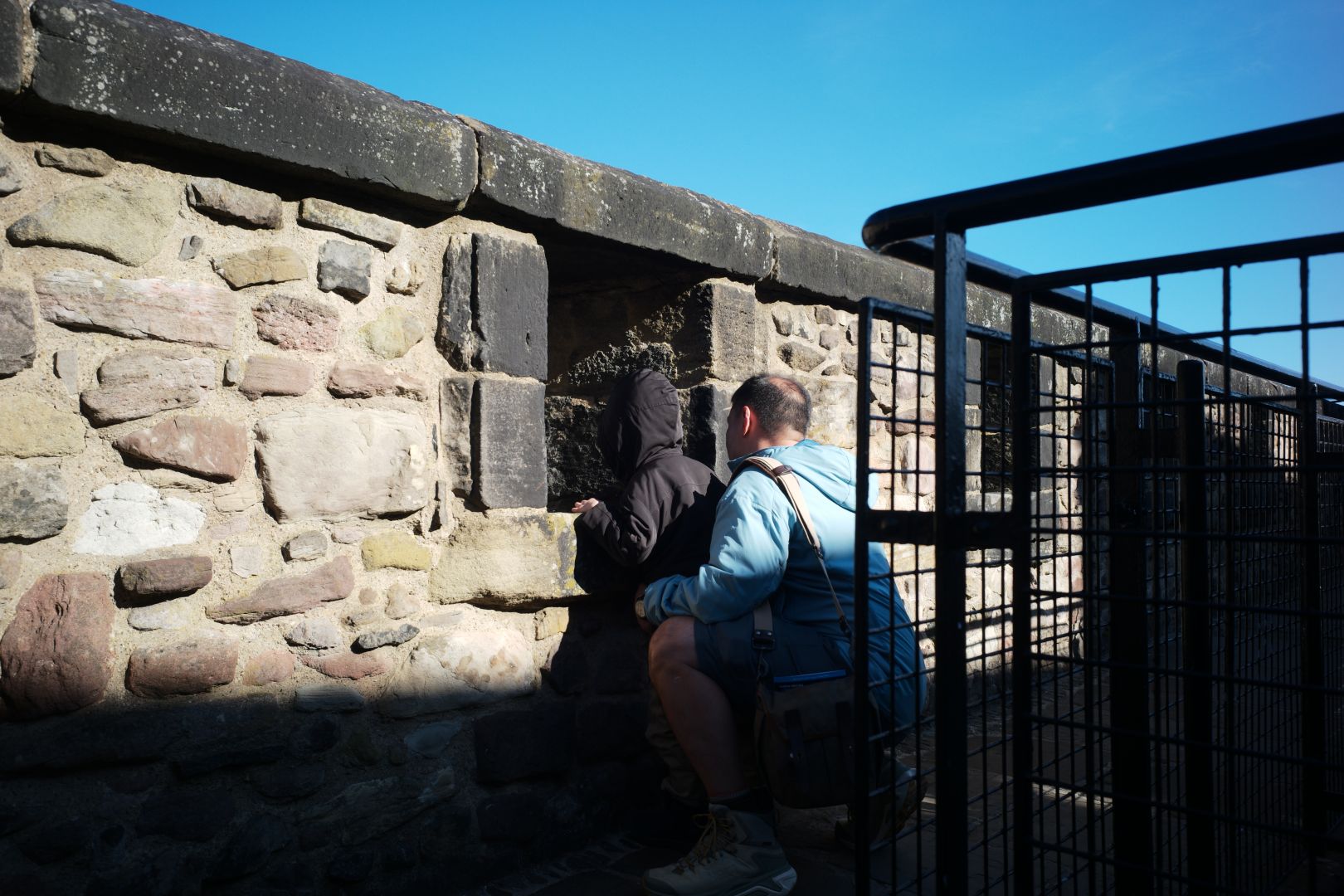 A young boy in a hooded black jacket and a man in blue huddled together to peer through an opening in the castle wall