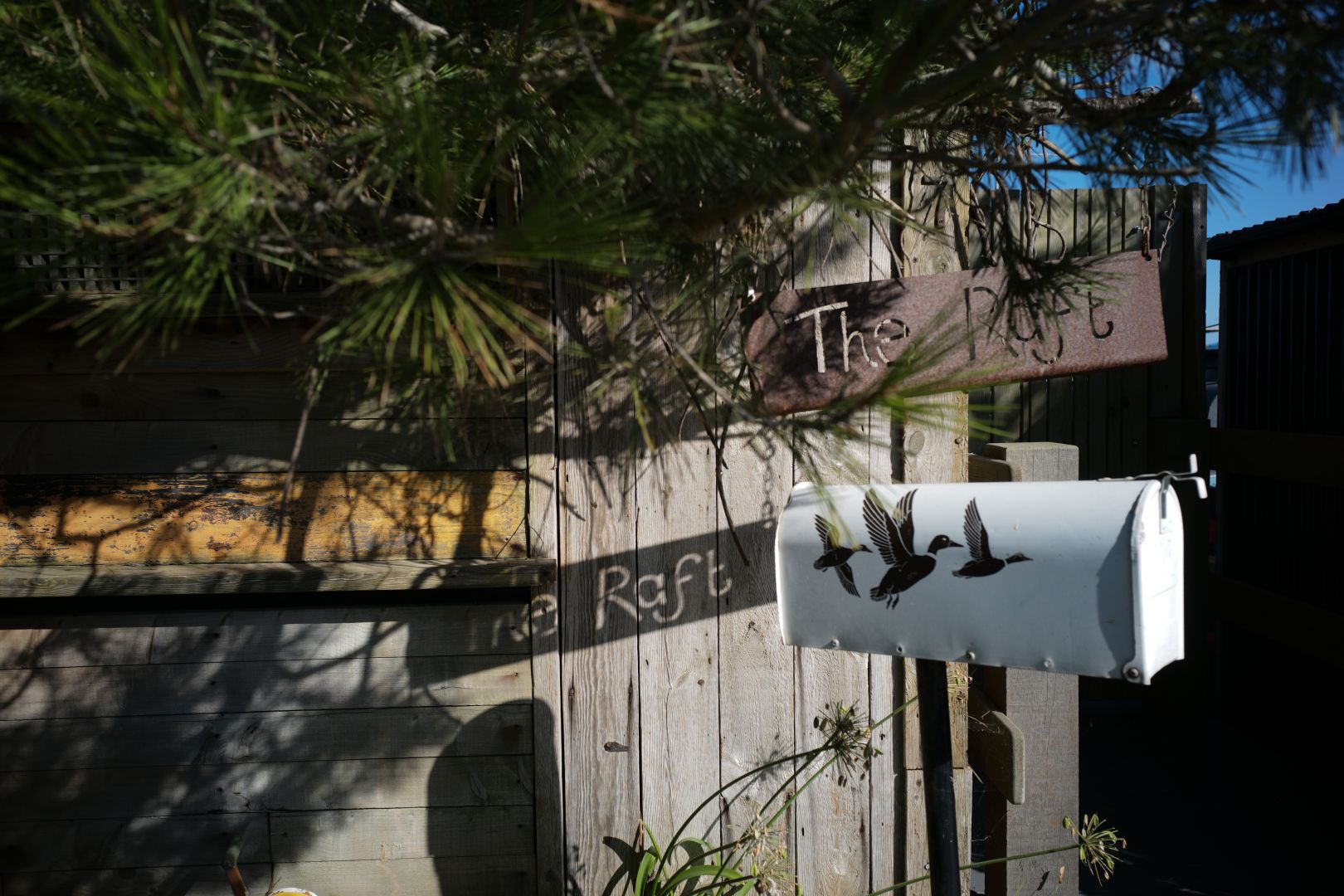 A boat house entrance. The boat is called The Raft, and its laser cut sigh is casting a shadow on its fence. Also pictured, a white mailbox with ducks, wings-spread, on them. A tree leans over the fence