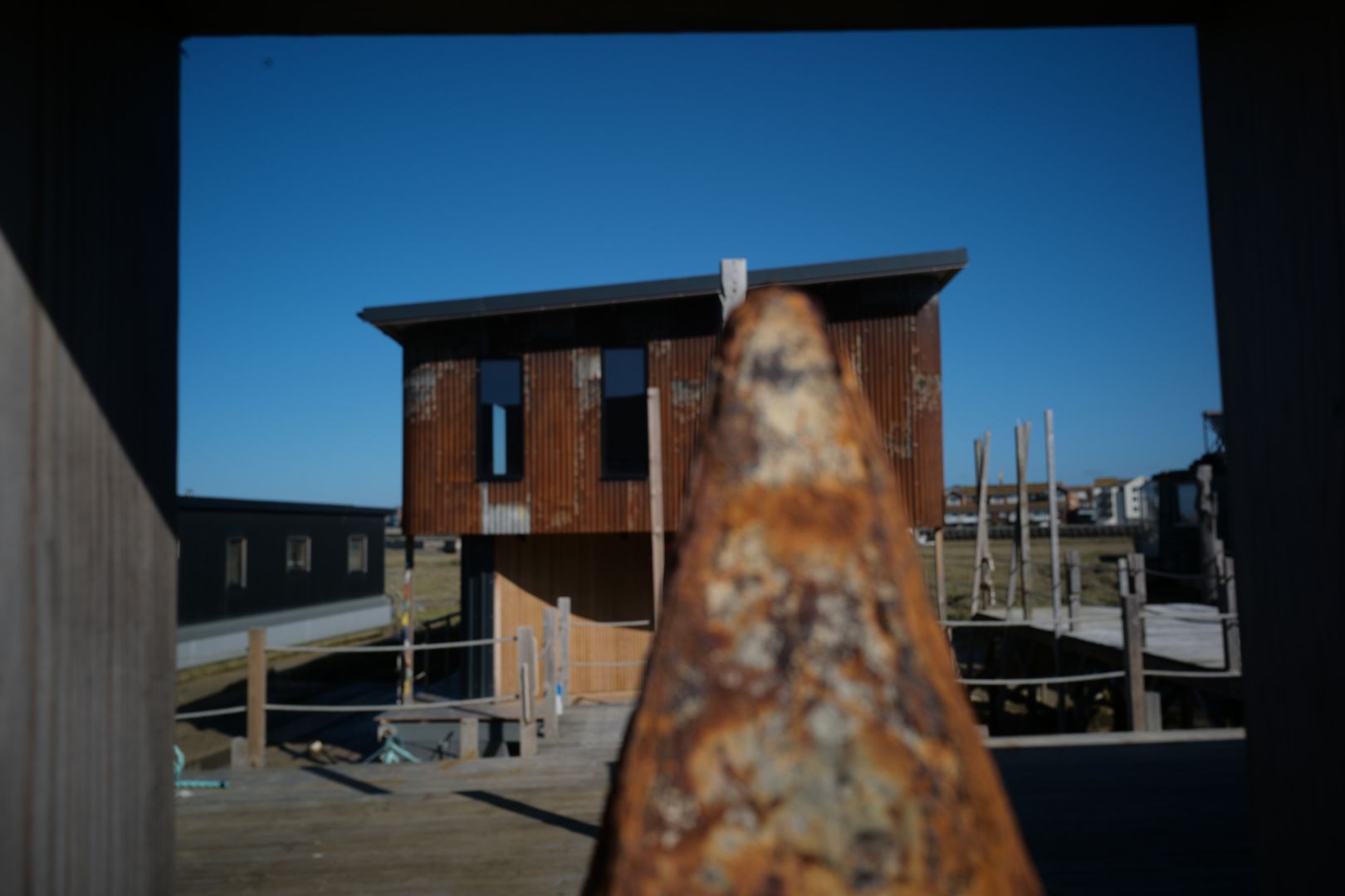 Picture taken through a gate of a boat house. In the front right-center is rusted meal beam partially obstructing the compound of the boathouse behind.