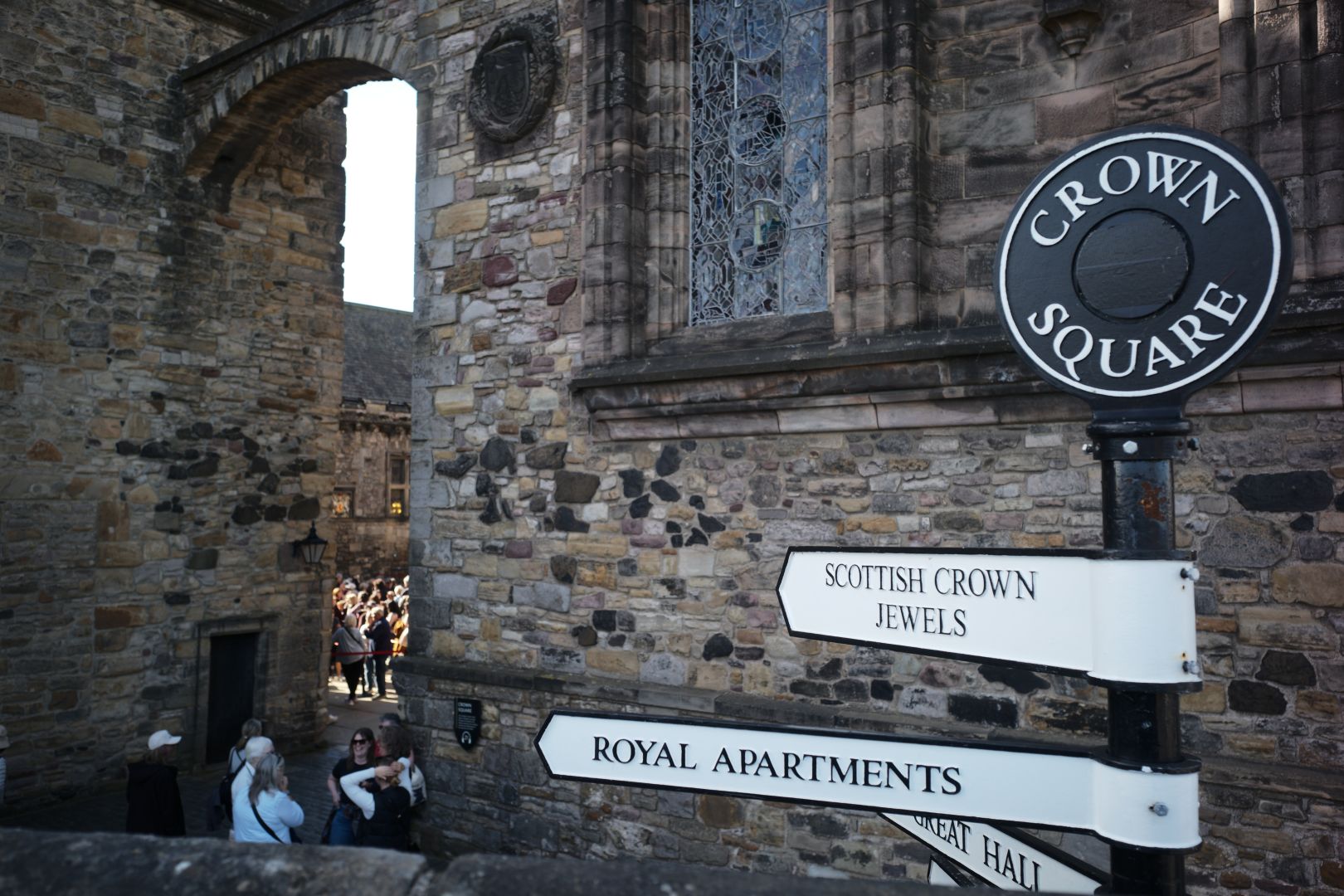 Castle signs pointing to SCOTTISH CROWN JEWELS, ROYAL APARTMENTS, (partially obscured) GREAT HALL. The top of the sign post is topped by a round disc which says “CROWN SQUARE”. Behind is a castle wall an entrance towards the square which the arrows point to.